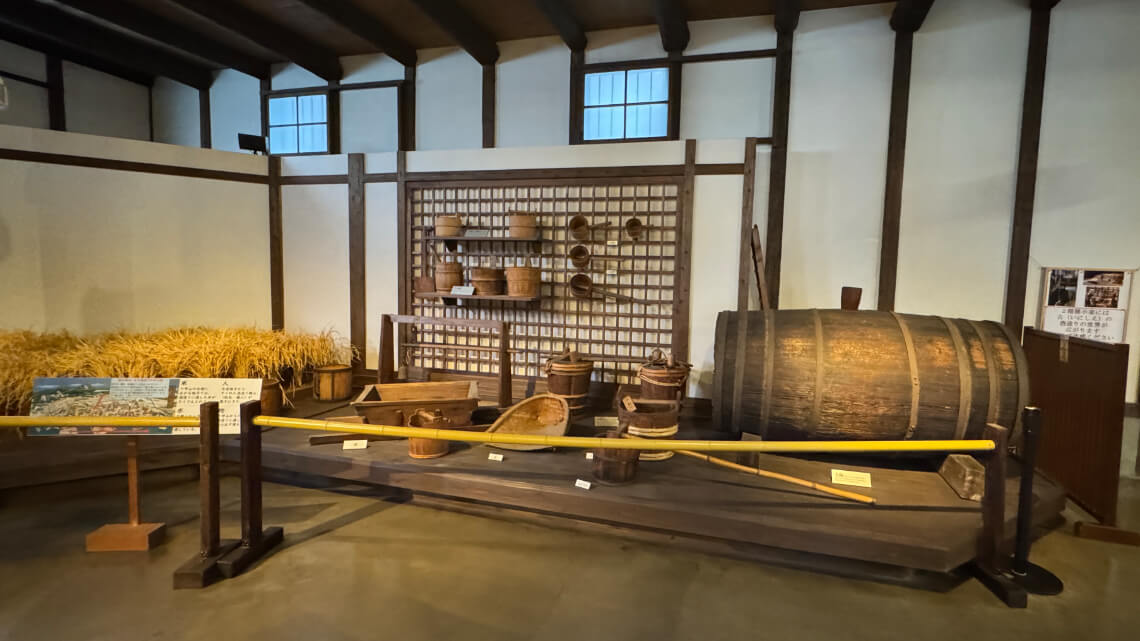 Display of traditional sake-making tools at Hakutsuru Sake Brewery Museum, including barrels and baskets, with a low yellow railing allowing easy viewing from a wheelchair.