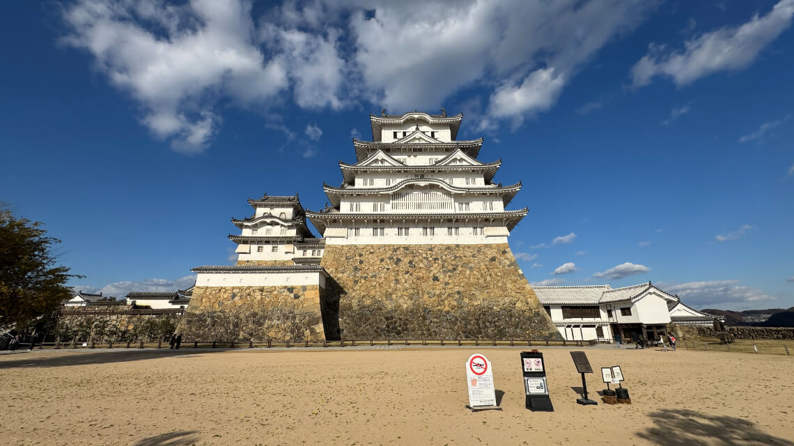 A stunning view of the castle keep from Bizenmaru Bailey, showcasing its towering white walls, stone base, and surrounding open courtyard.