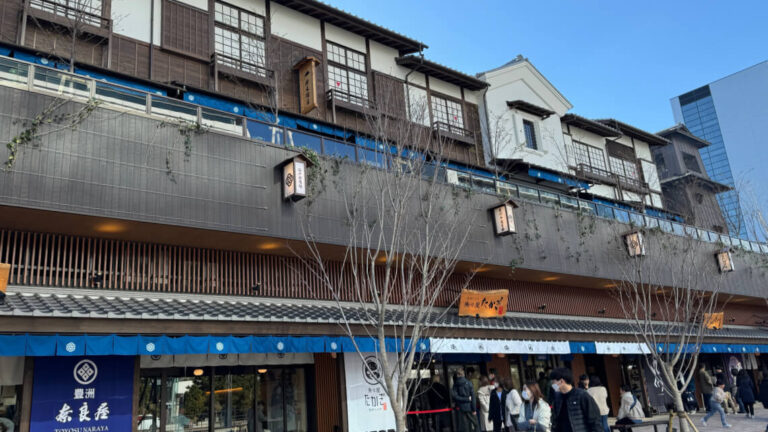 A traditional-style two-story building at Toyosu Senkyaku Banrai in Tokyo, featuring wooden facades, tiled roofs, and paper lanterns with Japanese text. The ground floor has blue fabric awnings with logos, shops, and people walking or standing in line.