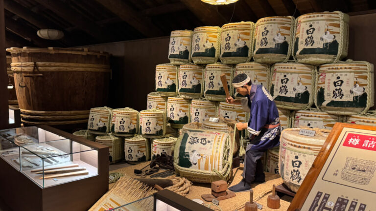 Mannequin in traditional attire working near sake barrels at Hakutsuru Sake Brewery Museum. Display cases show brewing tools.