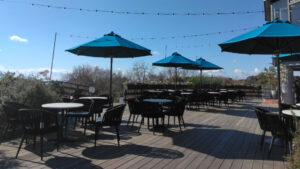 Outdoor rest area at the Glasshouse with tables, chairs, and blue umbrellas on a wooden deck.