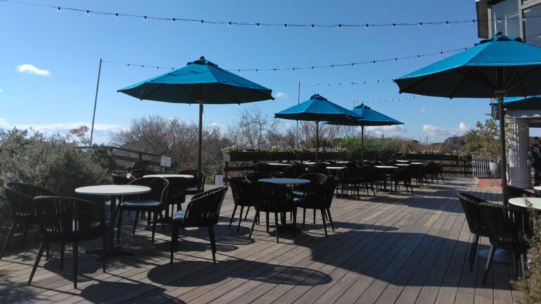 Outdoor rest area at the Glasshouse with tables, chairs, and blue umbrellas on a wooden deck.