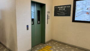 Elevator at the ropeway station with a green door, tactile paving, and a sign indicating access to the bottom station.