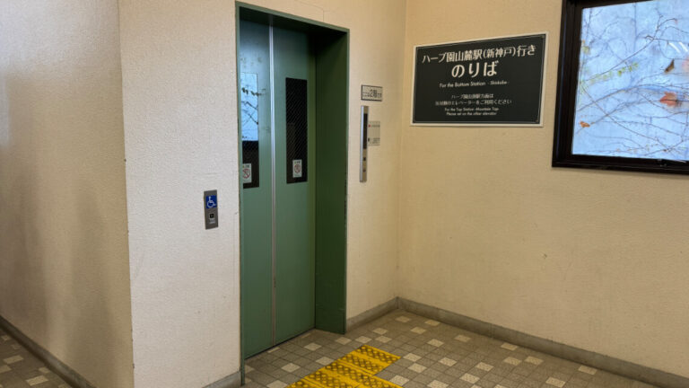 Elevator at the ropeway station with a green door, tactile paving, and a sign indicating access to the bottom station.