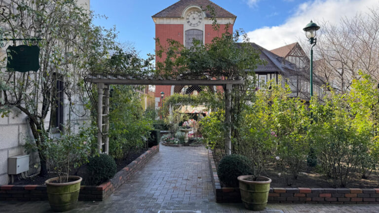 Pathway at Kobe Nunobiki Herb Garden, with greenery, flowerbeds, a wooden archway, and a clock-tower building in view.