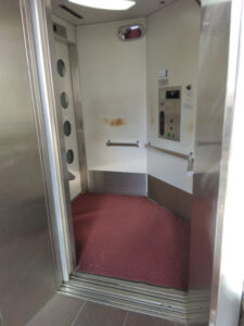 Interior of the elevator with low-positioned buttons, a red carpeted floor, handrails, and an exit through the opposite door.
