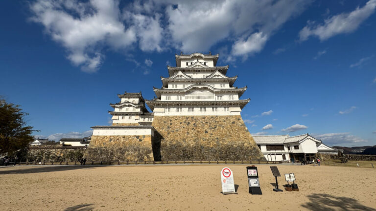 A view of the castle keep from Bizenmaru bailey, showcasing its towering white walls, stone base, and surrounding open courtyard.