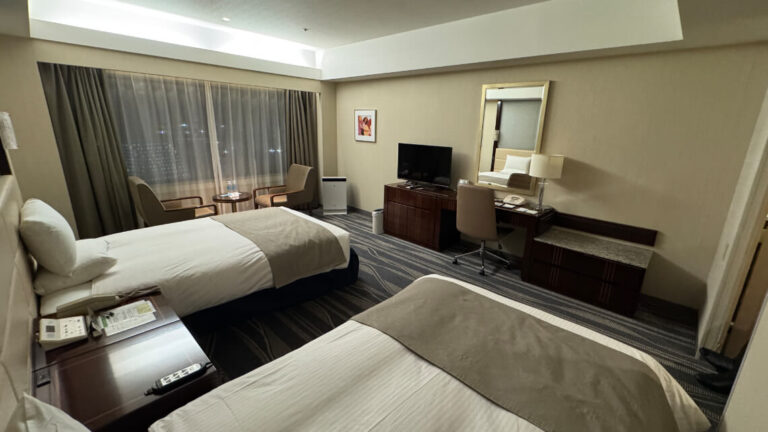 The accessible room includes two single beds, a desk with a chair, a TV on a low cabinet, a seating area by the window, and a wall-mounted mirror.
