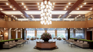 The lobby of Hotel Okura Kobe features a spacious layout with soft seating, a central floral arrangement, warm wood accents, and a large chandelier.