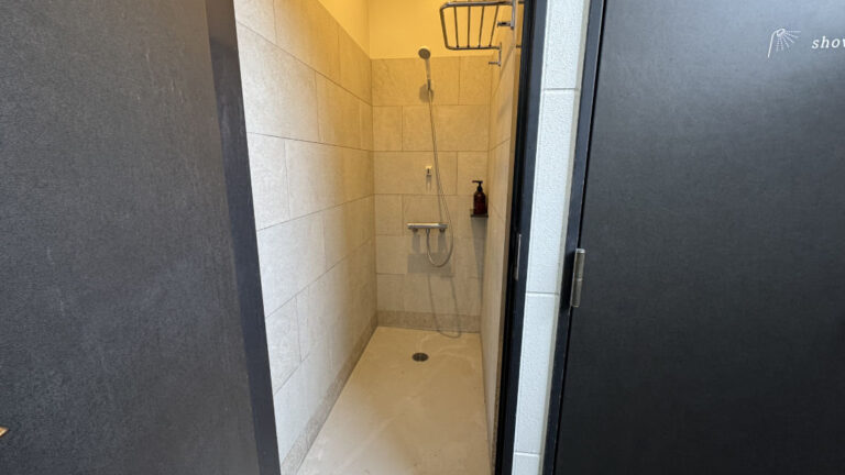 Open black door reveals a narrow tiled shower room with a handheld showerhead, shelf, and wall-mounted soap dispenser.