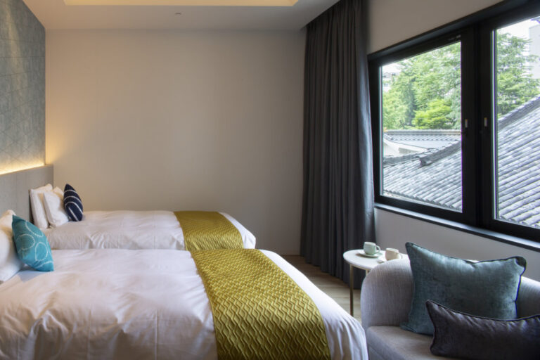Twin beds with green runners, a window-side seat, and a view of traditional tiled rooftops and trees outside.