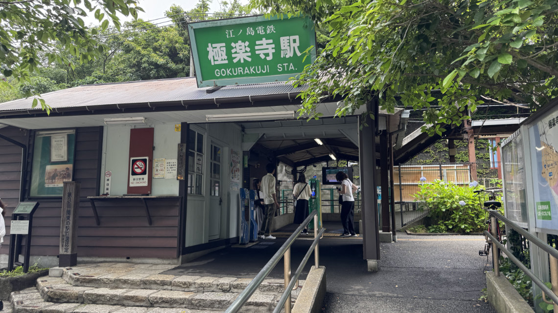 Gokurakuji Station, a small wooden building with a green sign and shaded by trees.