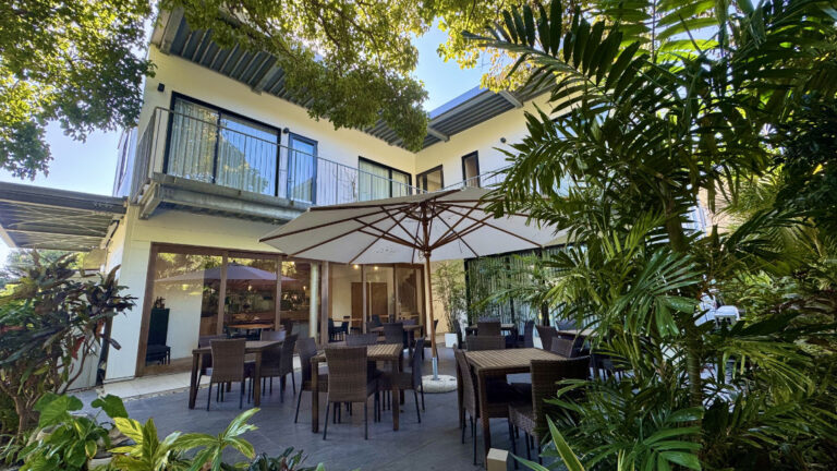 Daytime courtyard with wicker tables, large umbrella, lush greenery, and rooms with glass balconies above.