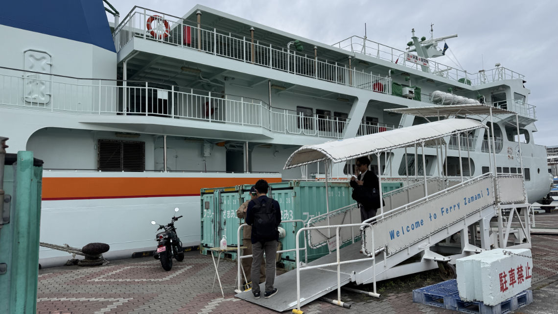 People boarding the Ferry Zamami 3 via a covered ramp, with containers and a parked motorcycle nearby on the dock.