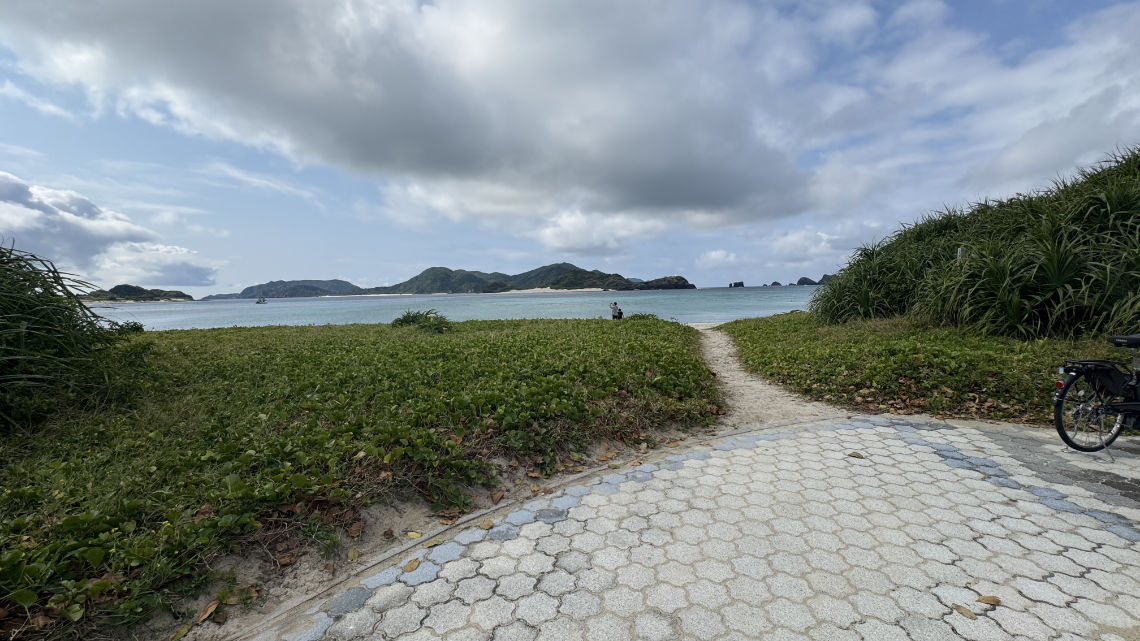 Paved path leading to a beach with lush greenery, calm blue sea, distant islands, and a person standing near the shoreline.