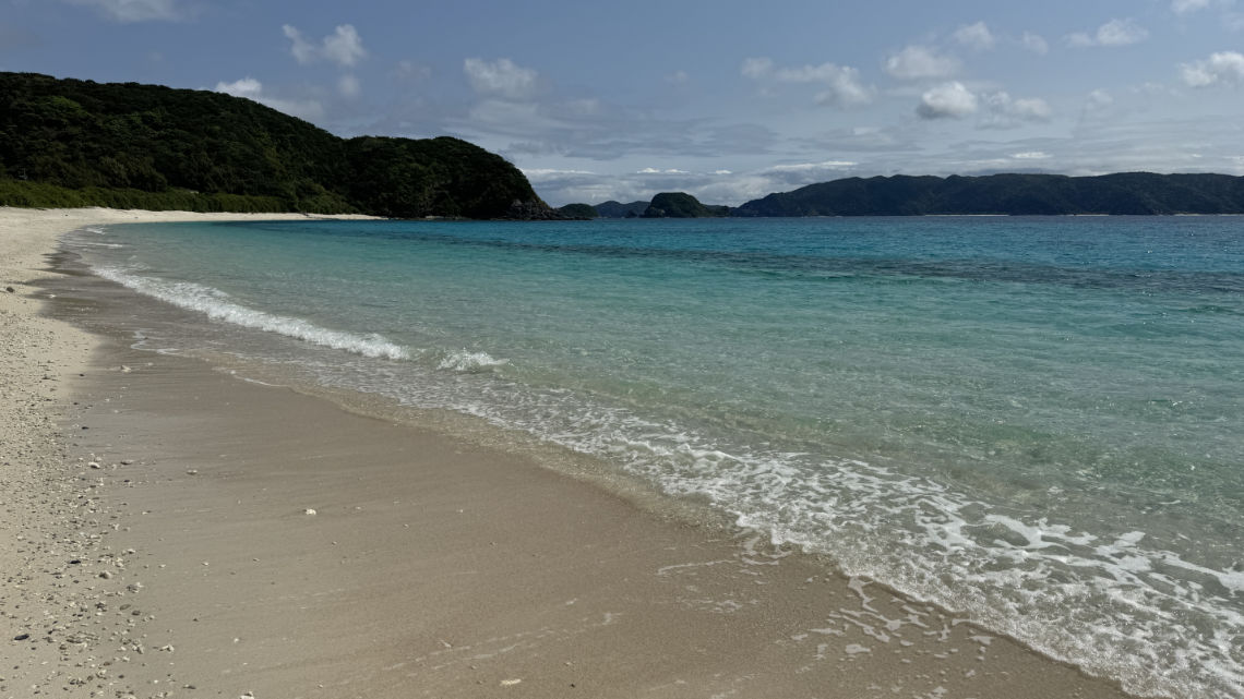 A quiet beach with soft sand and clear turquoise water, bordered by green hills and distant islands under a partly cloudy sky.